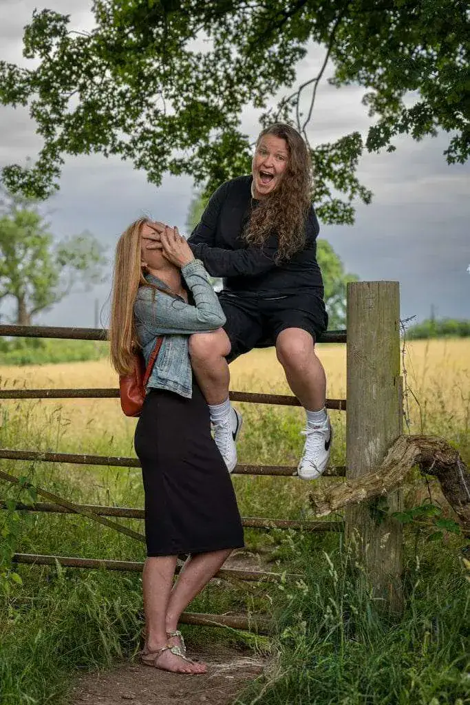 Two people enjoying a moment outdoors. One person, wearing a denim jacket and black skirt, stands laughing with a hand covering their mouth. The other, in a black outfit and white shoes, is sitting on a wooden fence, smiling widely. Image by David Kinsey Photography.