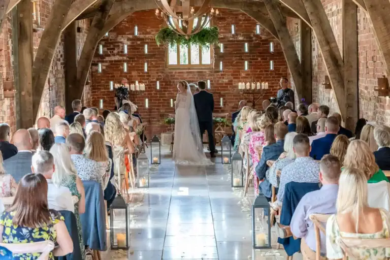 A bride and groom stand at the altar in a rustic brick venue with arched wooden beams. Guests are seated on both sides, and candles line the aisle. Sunlight filters through a window above the couple, creating a warm and intimate atmosphere. Image by David Kinsey Photography.