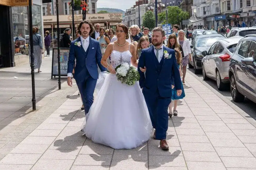 A bride in a white gown and two men in suits walk down a city street. The groom is on her right and a groomsman on her left. They're followed by a small group of people amidst parked cars and shops, under a clear blue sky. Image by David Kinsey Photography.