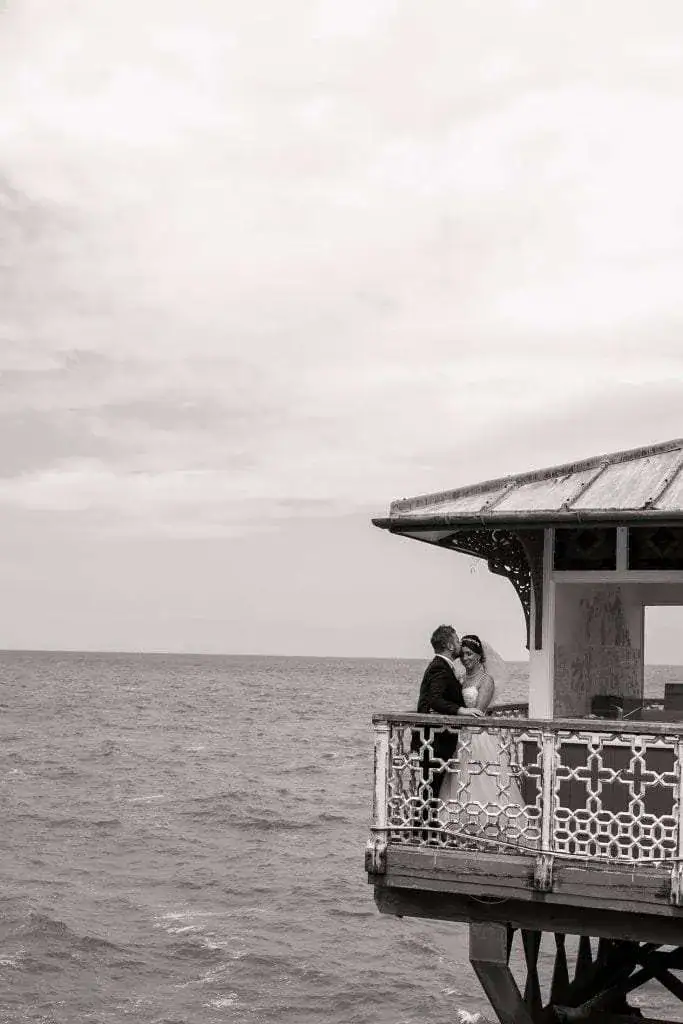 A black and white photo of a couple standing on a decorative balcony overlooking a vast ocean. The sky is partly cloudy, creating a serene atmosphere. The couple is embracing, gazing at each other affectionately. Image by David Kinsey Photography.