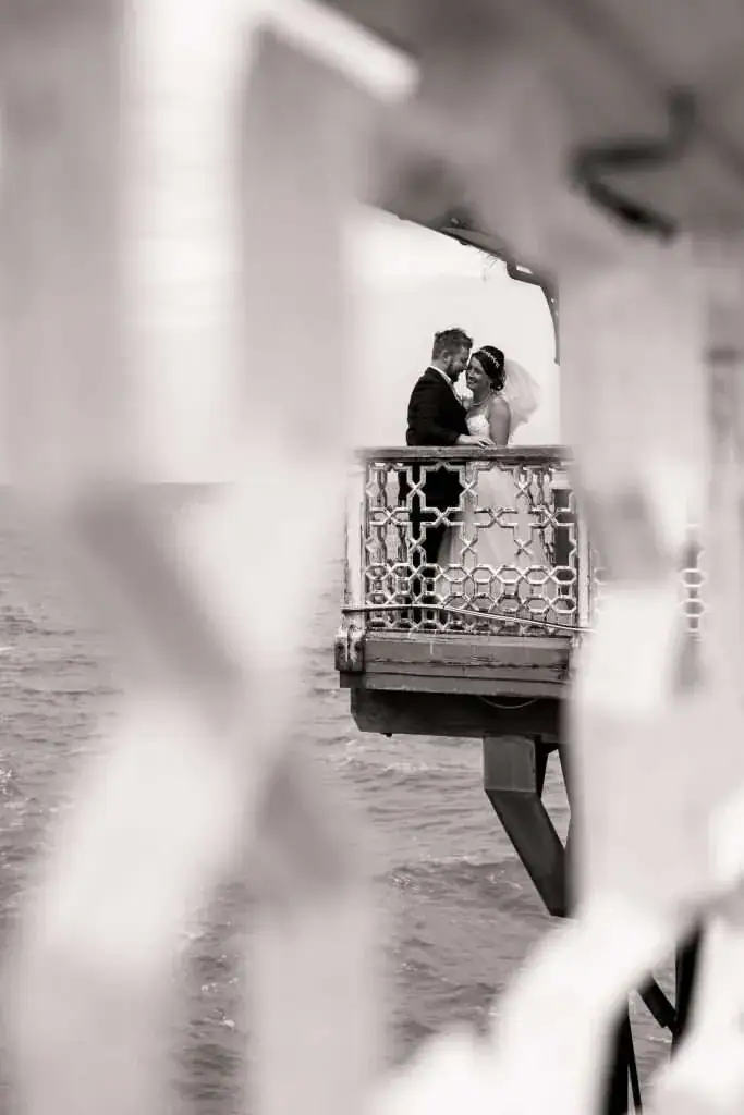 A bride and groom embrace on a balcony overlooking the sea. The image is framed by an out-of-focus railing in the foreground, creating a romantic and intimate scene. The photo is in black and white. Image by David Kinsey Photography.