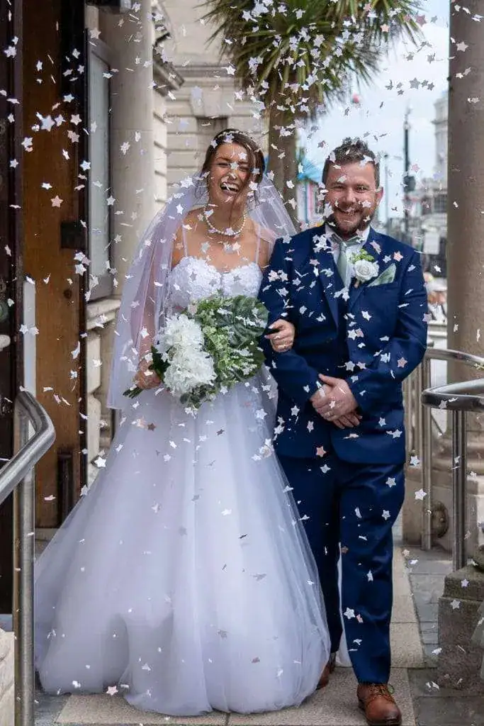 A bride and groom are walking down steps, smiling and laughing as they are showered with confetti. The bride is in a white gown holding a bouquet, and the groom is in a navy blue suit with a bow tie. A palm tree is in the background. Image by David Kinsey Photography.