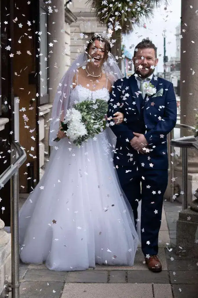A joyful bride and groom exit a building, arm in arm, smiling broadly. The bride wears a white gown and holds a bouquet of white flowers, while confetti falls around them. The groom is in a navy suit with a white boutonniere. Image by David Kinsey Photography.