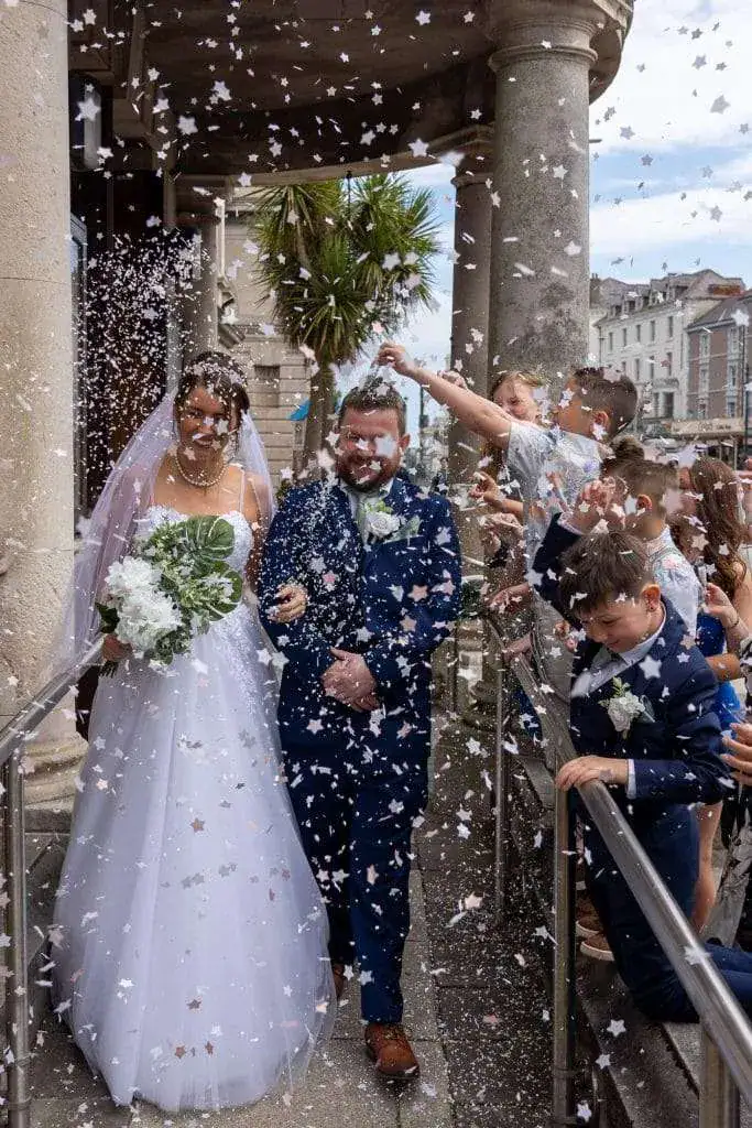 A bride in a white gown and a groom in a blue suit walk arm in arm while guests shower them with confetti. They are smiling and surrounded by friends and family celebrating outside a building. Image by David Kinsey Photography.