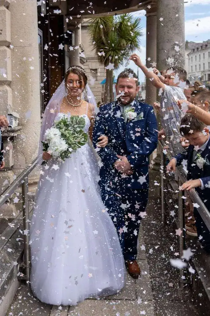 A bride and groom in wedding attire walk down steps outside a building, smiling as guests shower them with confetti. The bride holds a bouquet of white flowers. The scene is lively and full of celebration. Image by David Kinsey Photography.