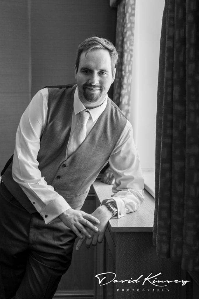 Black and white photo of a man in formal attire leaning against a wooden surface near a window. He wears a vest, shirt, tie, and watch, smiling slightly. Curtains and a textured wall are in the background. Image by David Kinsey Photography.