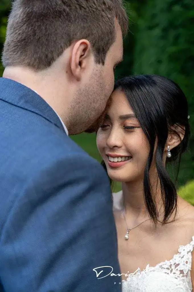 A couple embraces outdoors. The man in a blue suit gently kisses the forehead of a woman in a white lace dress, who is smiling with her eyes closed. The background is filled with greenery. Image by David Kinsey Photography.