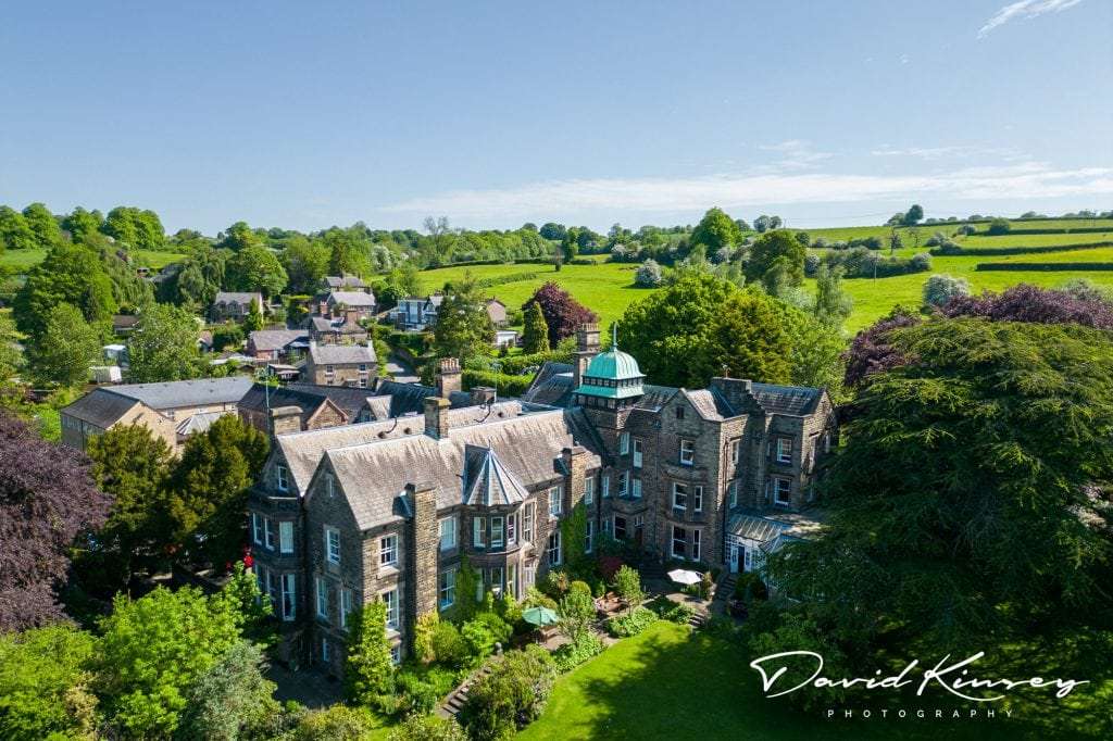 Aerial view of a large, historic stone mansion surrounded by lush green fields and trees under a clear blue sky. Nearby are smaller buildings and a small village, creating a picturesque countryside scene. Image by David Kinsey Photography.