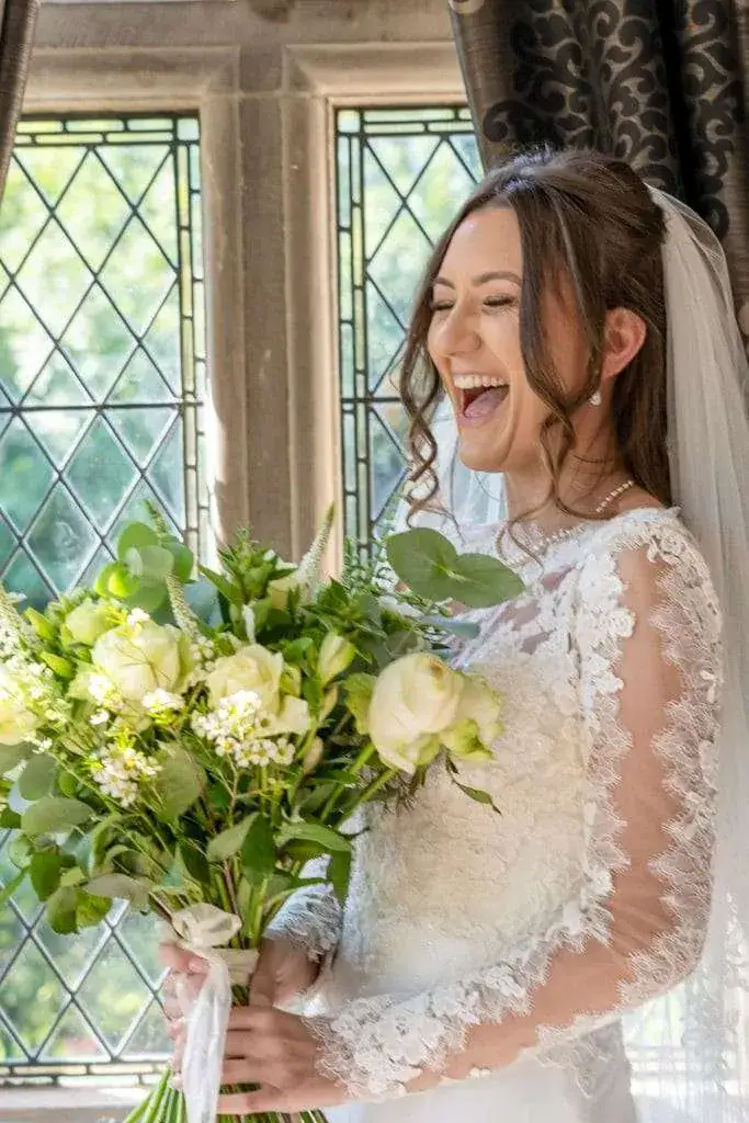 A joyful bride in a white lace gown holds a bouquet of white roses and greenery. She stands near a window with diamond-patterned glass, laughing. Sunlight filters through the window, illuminating the scene. Image by David Kinsey Photography.