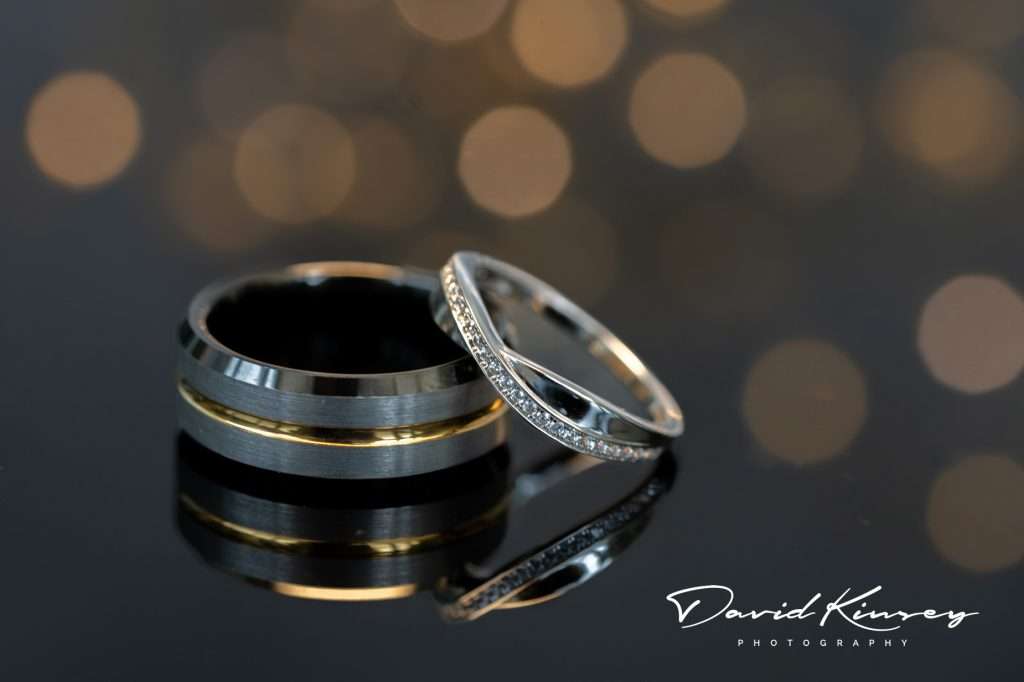 Makeney Hall Wedding Photographer. Wedding rings on shiny black surface with reflection and blurred fairy lights in the background