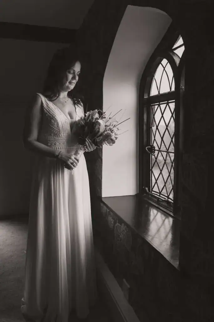A bride in a long, elegant gown stands by a stained glass window, holding a bouquet. She looks down, illuminated by soft natural light filtering through the window. The scene is in black and white, creating a timeless, serene atmosphere. Image by David Kinsey Photography.