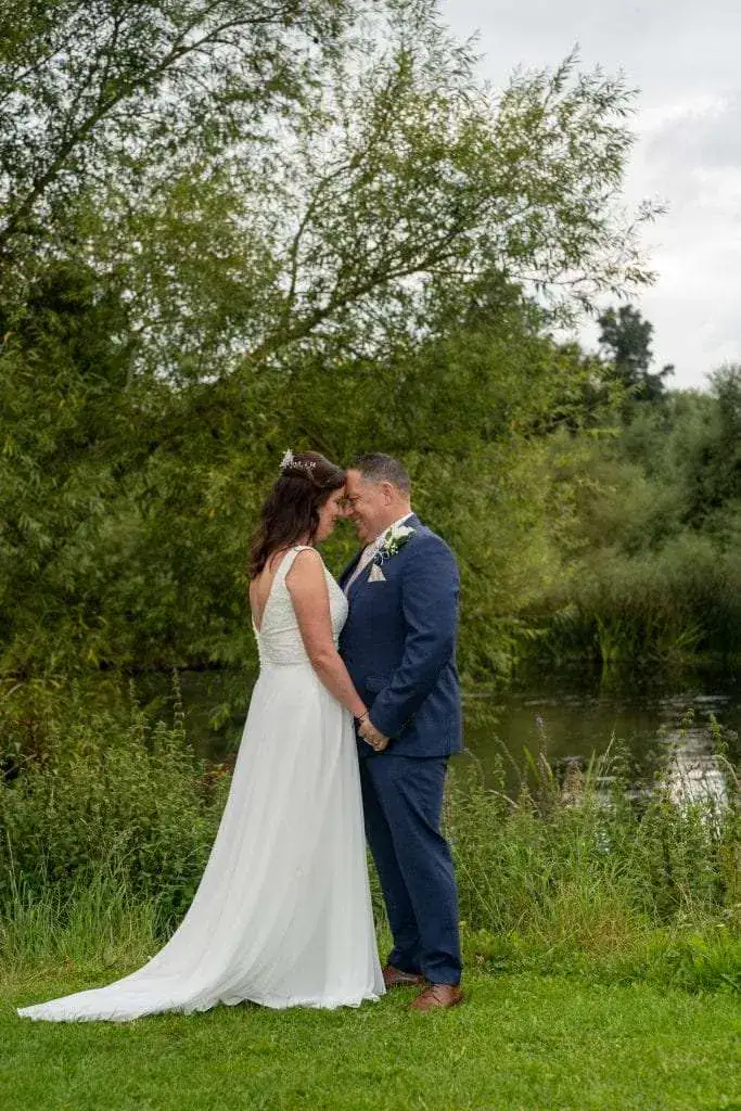 A bride in a white dress and a groom in a blue suit stand closely together, holding hands, with their foreheads touching. They are outdoors, surrounded by lush greenery near a body of water. Image by David Kinsey Photography.