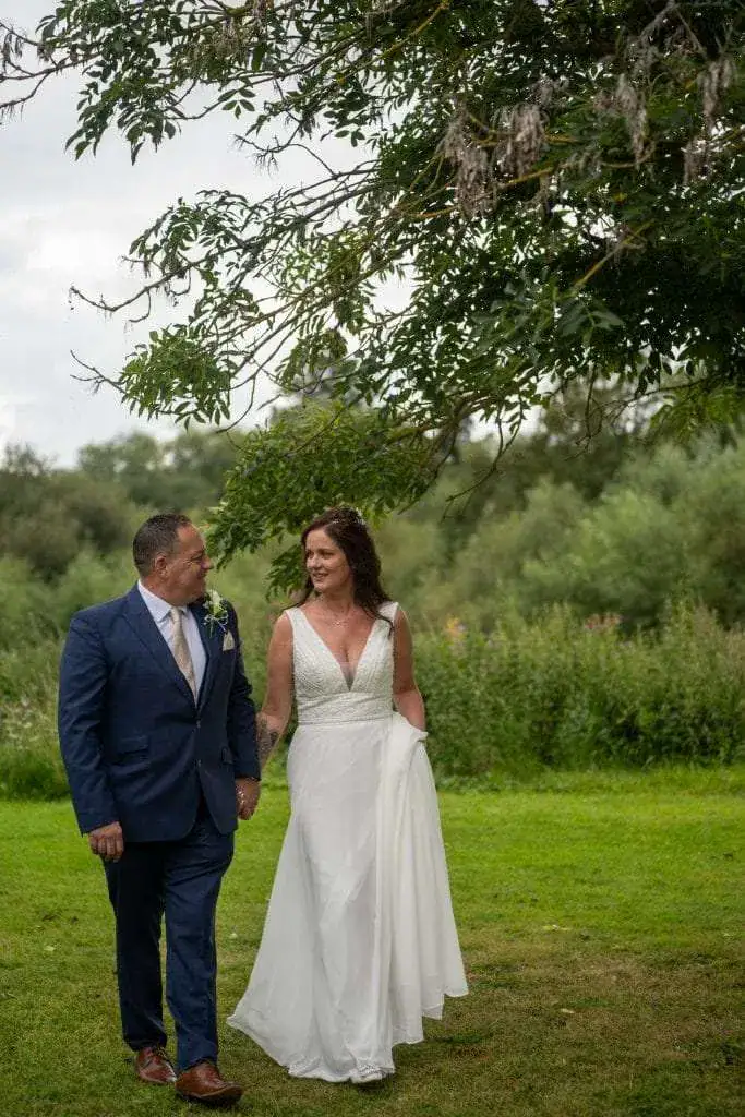 A couple walks on a lush green lawn under a tree. The woman is in a white wedding dress, holding her skirt, while the man wears a blue suit. They are looking at each other, smiling, with greenery in the background. Image by David Kinsey Photography.