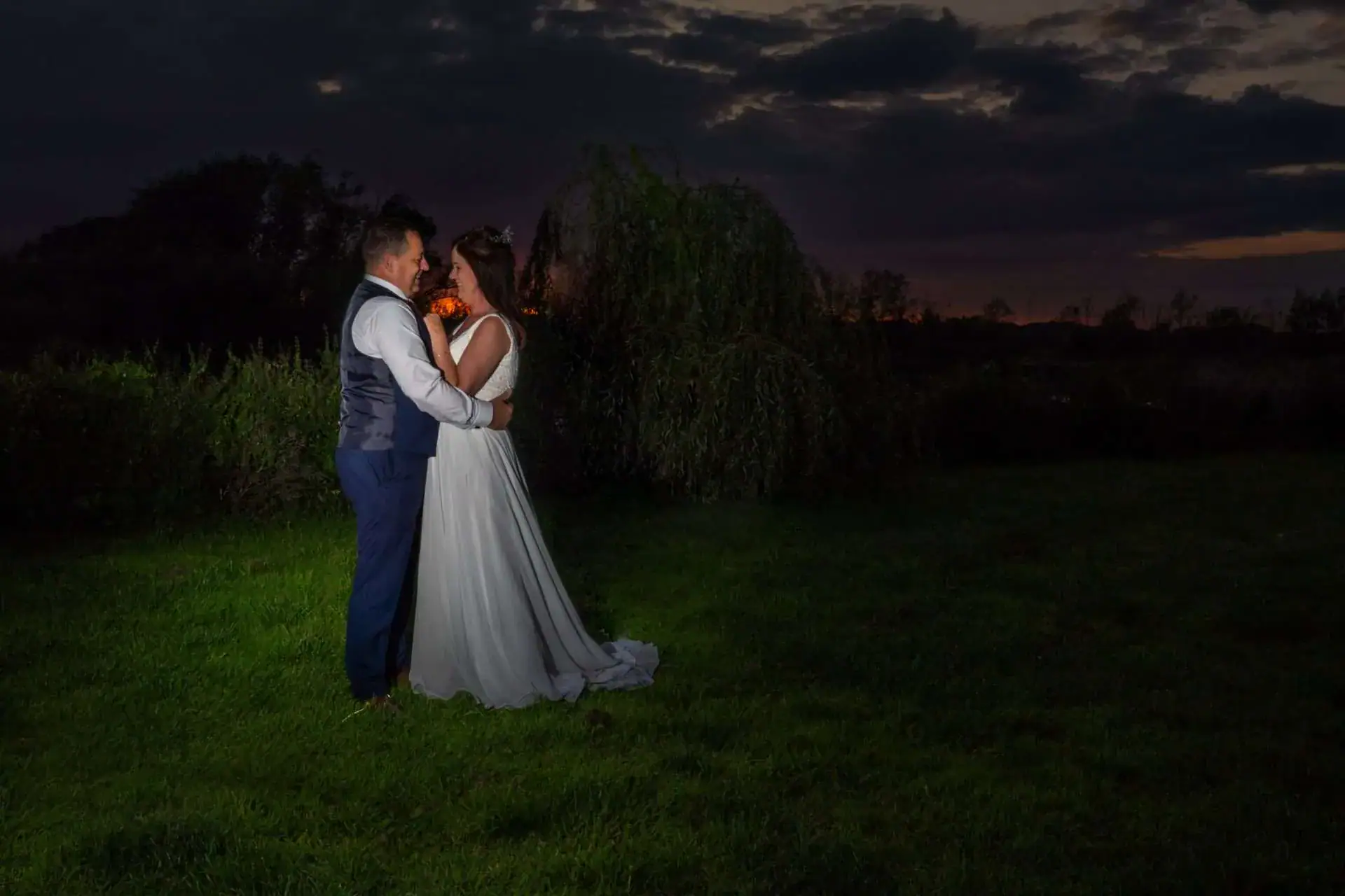 A couple in wedding attire stands closely together on a grassy field at dusk, under a dark, cloudy sky. They are surrounded by greenery, with a faint hint of sunset on the horizon, creating a romantic and serene atmosphere. Image by David Kinsey Photography.